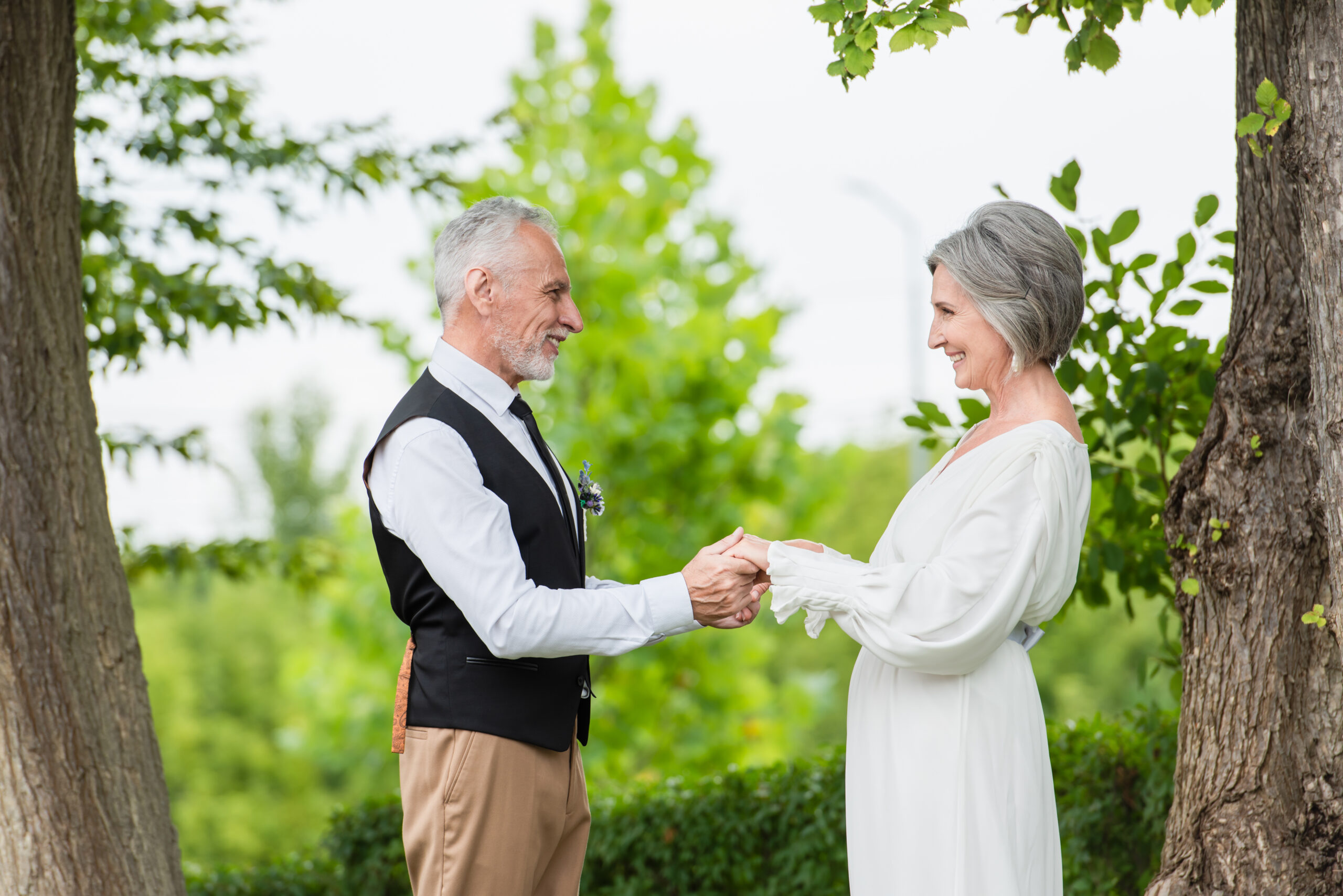 side view of cheerful mature man in formal wear holding hands with smiling bride in wedding dress in